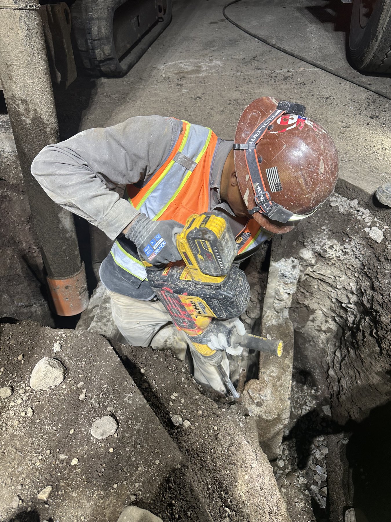 DeWalt 60V cordless SDS Max rotary hammer with 20V battery being used to break concrete around utility pipe in a deep trench, worker in hi-vis and hard hat