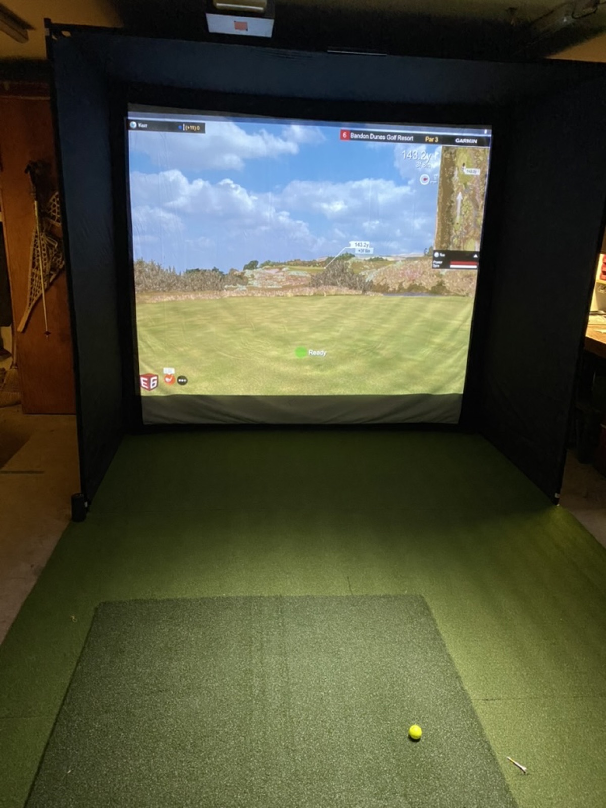 V1 garage golf simulator on Dec 13 2022 - black-walled Net Return enclosure framing a wall impact screen rendering the Bandon Dunes Par 3 hole with Garmin R10 tracking, putting green mat on the garage floor with hitting strip and tee box visible