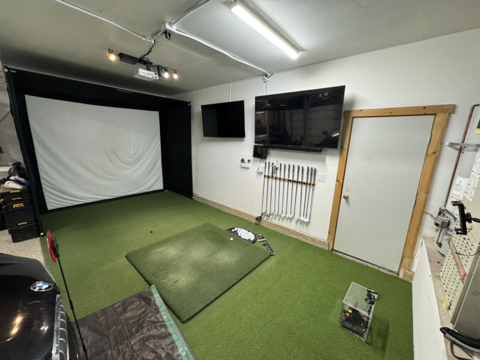 V2 garage golf simulator on May 26 2024 - built-out garage bay with permanent ceiling-mount short-throw projector under track lights, large wall-anchored impact screen, dual TV monitors mounted on side wall, wall-mounted club rack with full set, wall-to-wall green putting carpet, Garmin R10 on stand at the tee box, BMW SUV visible at left in same garage