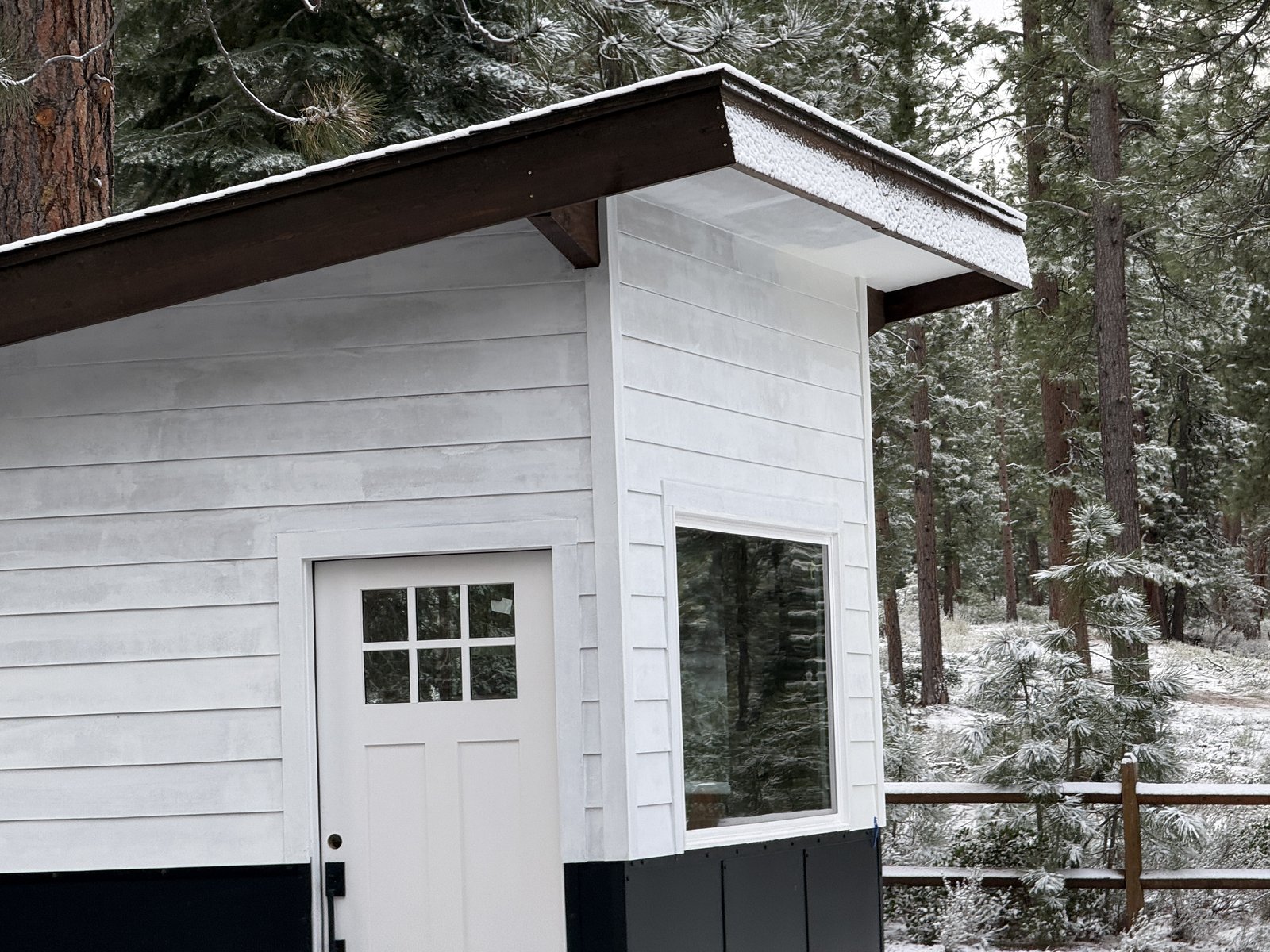 Royal Way Shed finished reveal — 10×12 mono-slope cream lap siding with matte-black wainscot, JW casement window, dark metal roof, dusting of snow on the roof, surrounded by Truckee pines, April 2026
